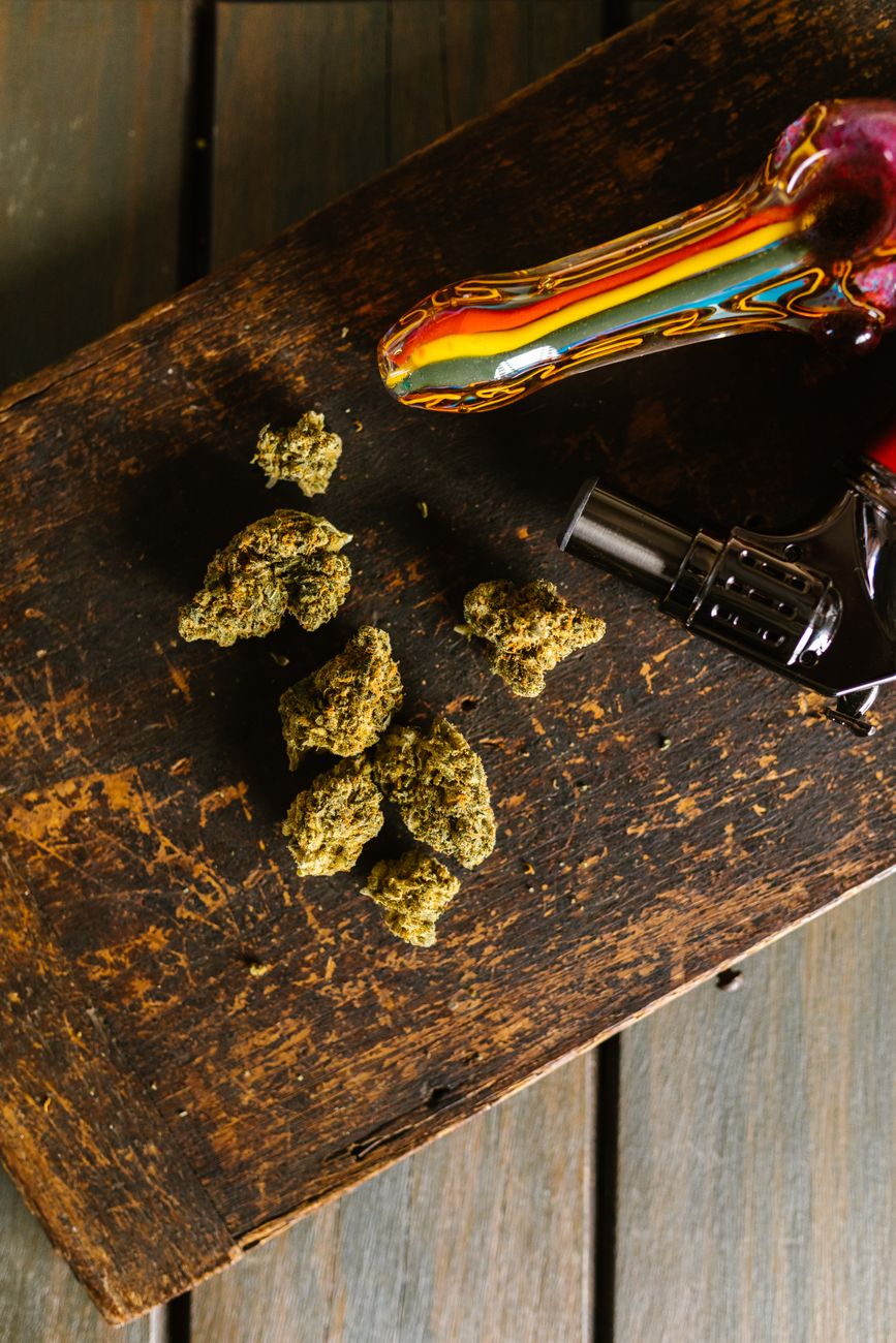 Eine bunte Glaspfeife und Cannabisblüten auf einem rustikalen Holztisch, Hochformat.
