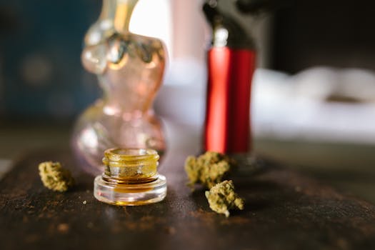 Glas mit Cannabiskonzentrat, umgeben von getrockneten Marihuanablüten und Zubehör auf einem Tisch.
